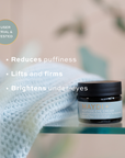 Maydi+ eye cream jar with product benefits text on a blurred background. reduces puffiness, lifts and firms, brightens under-eyes.